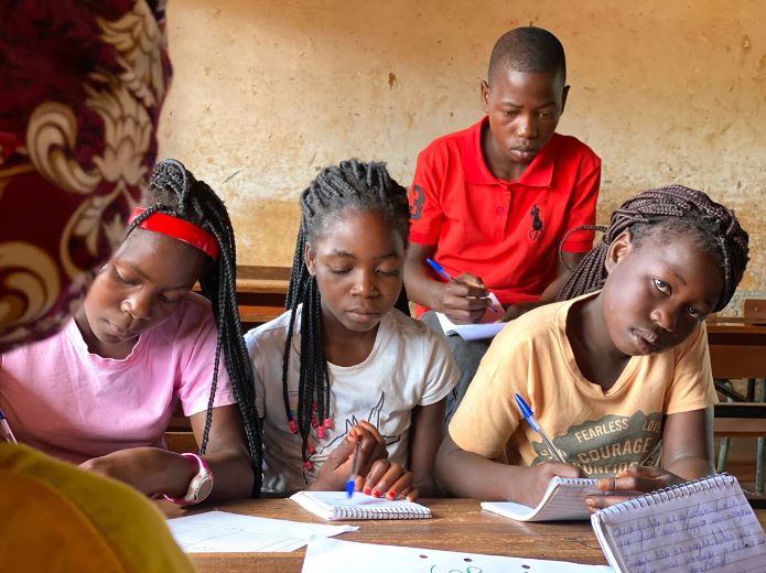 4 jovens num ambiente de escola escrevem em pequenos blocos de linhas com canetas. Uma delas olha em direção à câmara.