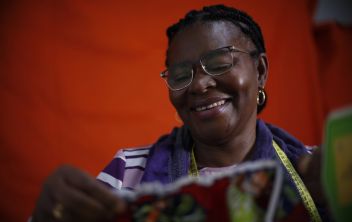 Mulher sorridente a olhar para baixo, na direção da peça de roupa que acabou de costurar e segura com as mãos, com uma fita métrica de costura ao pescoço e uma máquina de costura ao lado, num fundo vermelho.