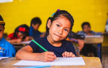 Menina com uma t-shirt azul, num ambiente interior de escola, olha diretamente para a câmara enquanto segura um lápis de cor verde para desenhar numa folha branca.