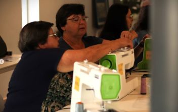 Duas senhoras, sentadas frente a duas máquinas de costura, enquanto uma delas ajuda a outra na preparação da máquina para costurar, num ambiente de workshop de costura, do projeto Mulheres em Ação.