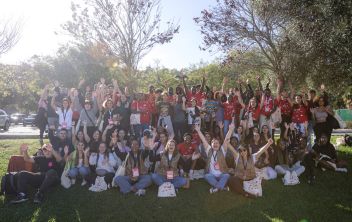 Fotografia de grupo tirada ao ar livre: dezenas de jovens e alguns formadores estão sentados e em pé num relvado, muitos com t-shirts vermelhas e sacos do 'Bora Jovens. Todos sorriem e acenam para a câmara, transmitindo um ambiente de celebração, união e participação no programa 'Bora Jovens, o programa de empregabilidade jovem da Ajuda em Ação em Portugal.