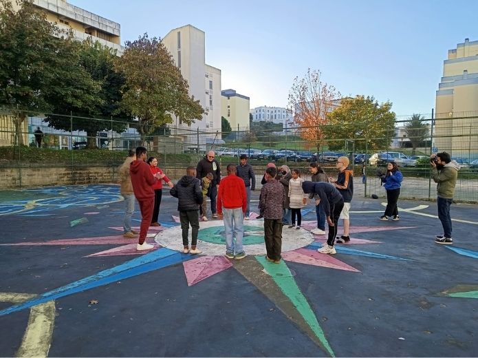 Um grupo diverso de jovens, em círculo, num parque de basquetebol ao ar livre, com vários desenhos coloridos no chão, incluindo o de uma bússola. O ambiente é de atenção, concentração e inclusão.
