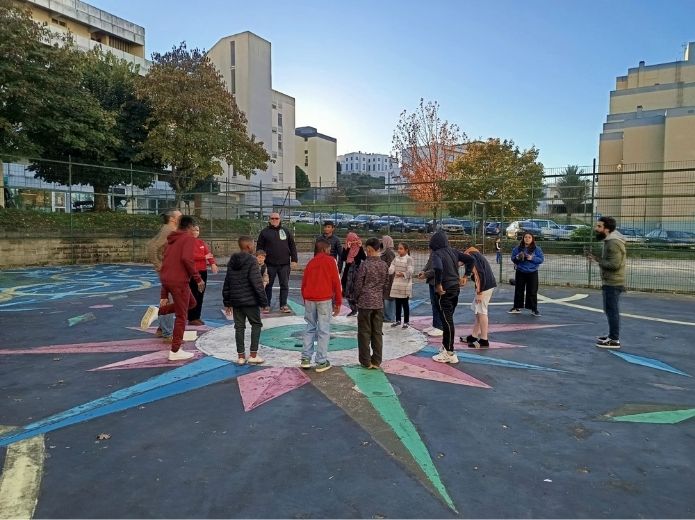 Um grupo diverso de jovens, em círculo, num parque de basquetebol ao ar livre, com vários desenhos coloridos no chão, incluindo o de uma bússola. O ambiente é de atenção, concentração e inclusão.