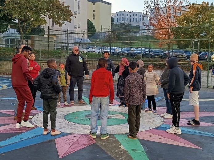 Um grupo diverso de jovens, em círculo, num parque de basquetebol ao ar livre, com vários desenhos coloridos no chão, incluindo o de uma bússola. O ambiente é de atenção, concentração e inclusão.