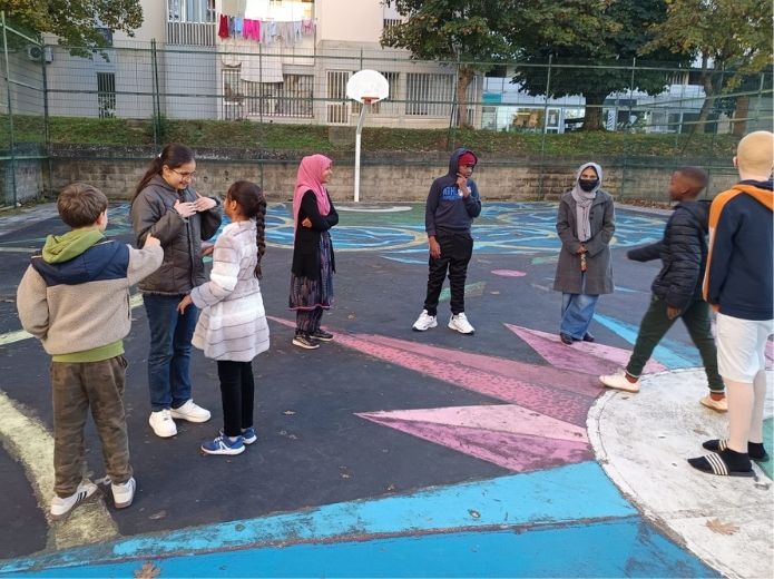 Um grupo diverso de jovens a interagir entre si, num parque de basquetebol ao ar livre, com vários desenhos coloridos no chão, incluindo o de uma bússola. O ambiente é de alegria, concentração e inclusão.