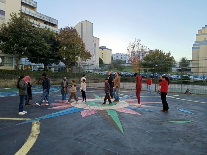Um grupo diverso de jovens, num parque de basquetebol ao ar livre, com vários desenhos coloridos no chão, incluindo o de uma bússola.
