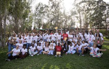 Fotografia de grupo tirada ao ar livre: dezenas de jovens e alguns formadores estão sentados e em pé num relvado, com t-shirts brancas do 'BORA Jovens. Todos sorriem e acenam para a câmara, transmitindo um ambiente de celebração, união e participação no programa 'BORA Jovens, o programa de empregabilidade jovem da Ajuda em Ação em Portugal.