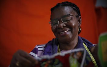 Mulher sorridente a olhar para baixo, na direção da peça de roupa que acabou de costurar e segura com as mãos, com uma fita métrica de costura ao pescoço e uma máquina de costura ao lado, num fundo vermelho.