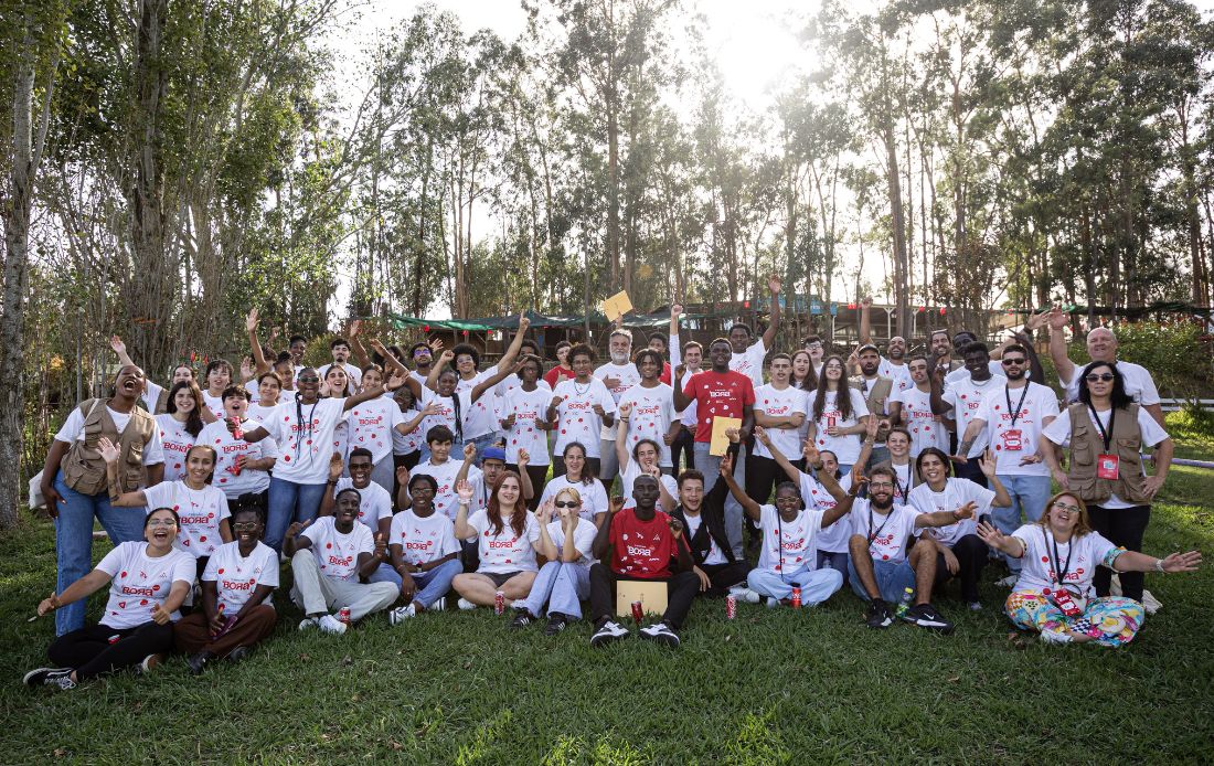 Fotografia de grupo tirada ao ar livre, na 4ª Edição do Bootcamp 'BORA Jovens, o programa de empregabilidade jovem da Ajuda em Ação em Portugal, onde dezenas de jovens sorriem e acenam para a câmara, transmitindo um ambiente de celebração.